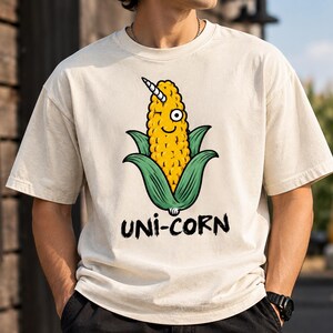May include: A cream-colored short-sleeve t-shirt featuring a graphic of a yellow corn cob with a white unicorn horn and a single eye. The corn is surrounded by green husks, and the text "UNI-CORN" is printed in black below the illustration.