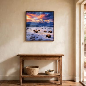 Square bison wall art framed above a wooden console table in a rustic entryway, winter buffalo herd and snow-capped mountains poster