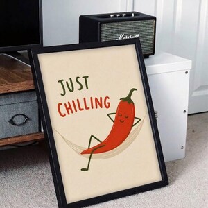 May include: A framed wall art print featuring a red chili pepper character relaxing in a hammock. The text "JUST CHILLING" is printed in green and red above the illustration. The artwork is set against a light cream background within a black frame.