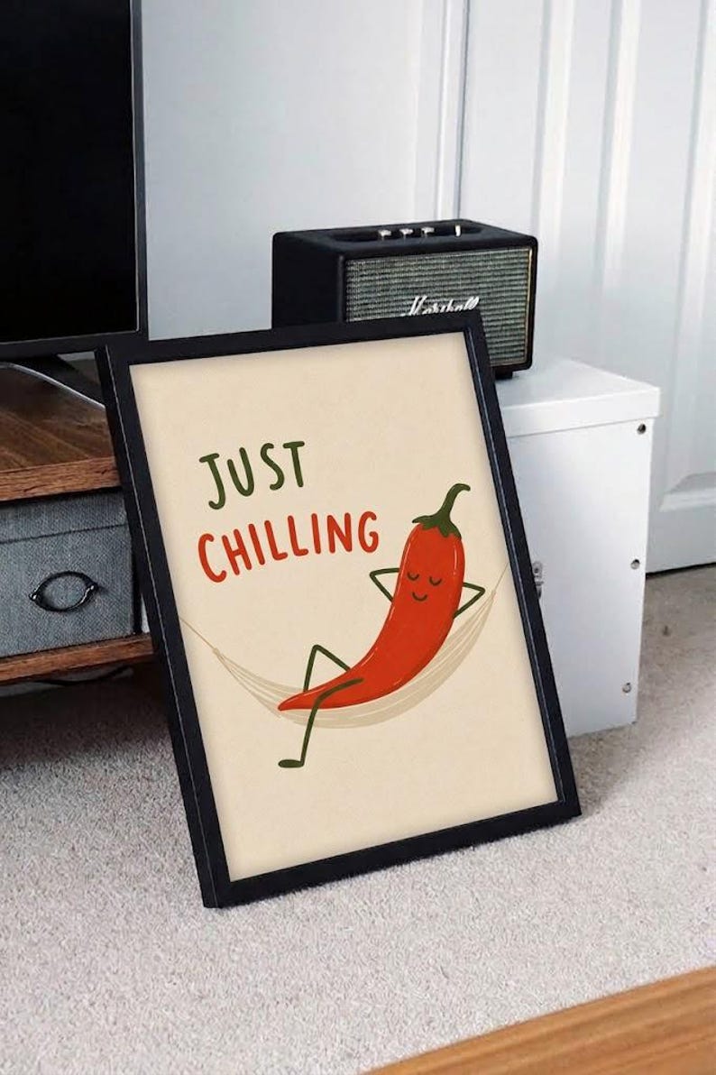 May include: A framed wall art print featuring a red chili pepper character relaxing in a hammock. The text "JUST CHILLING" is printed in green and red above the illustration. The artwork is set against a light cream background within a black frame.