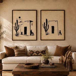 May include: Two framed wall art prints featuring black ink illustrations of desert scenes with cacti, adobe buildings, and figures in hats on a beige background, displayed side-by-side above a cream-colored sofa in a living room setting.