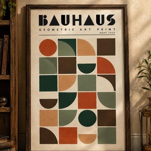 K&ouml;nnte beinhalten: Ein gerahmter Kunstdruck mit der Aufschrift BAUHAUS GEOMETRIC ART PRINT SEPT 1919. Das Poster zeigt ein Raster aus Quadraten mit verschiedenen geometrischen Formen wie Kreisen, Halbkreisen und Vollquadraten in Orange-, Gr&uuml;n- und Braunt&ouml;nen.