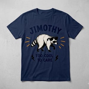 May include: A navy blue short-sleeve t-shirt featuring a graphic of a white raccoon wearing black sunglasses. The text JIMOTHY is printed above the raccoon, and the text TOO COOL TO CARE is printed below, flanked by two black lightning bolt icons.