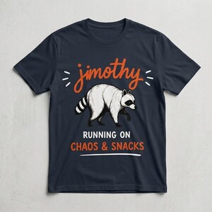 May include: A navy blue short-sleeve t-shirt featuring a graphic of a white raccoon. Above the raccoon is the orange text "jimothy" with small white dashes. Below the raccoon is the white text "RUNNING ON" and the orange text "CHAOS & SNACKS".