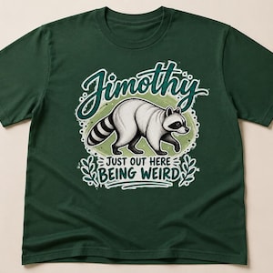 May include: A dark green short-sleeve t-shirt featuring a graphic of a white raccoon with black markings. The text "Jimothy" appears above the raccoon, and the text "JUST OUT HERE BEING WEIRD" is printed below, surrounded by decorative leaf accents.