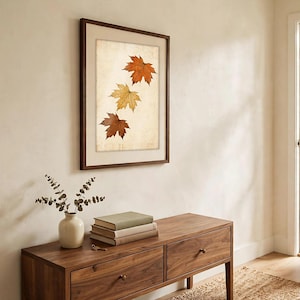 May include: A framed wall art piece featuring three autumn maple leaves in shades of orange, yellow, and brown, displayed vertically against a light, textured background within a dark wood frame.
