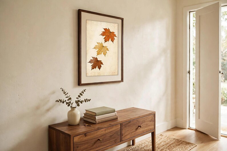 May include: A framed wall art piece featuring three autumn maple leaves in shades of orange, yellow, and brown, displayed vertically against a light, textured background within a dark wood frame.