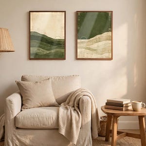 May include: Two rectangular wall art prints in wooden frames featuring abstract landscape designs with layered green and beige tones, each displaying a small white arched line graphic, displayed side by side on a plain wall.