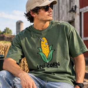 May include: An olive green short-sleeve t-shirt featuring a graphic of a yellow corn cob with a white unicorn horn, a single eye, and a small smile. Below the graphic, the text "UNI-CORN" is printed in a bold, black, hand-drawn style font.