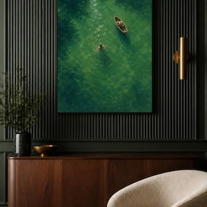 May include: A framed wall art print featuring an aerial view of a small wooden rowboat floating on vibrant green water with a person swimming nearby. The scene captures sunlight reflecting off the water surface, creating bright highlights in the sea.