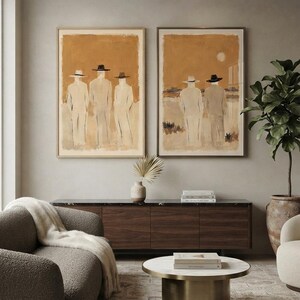 May include: Two rectangular framed wall art prints featuring abstract figures in hats against a warm ochre background. The left print displays three figures standing side by side, while the right print shows two figures in a landscape with a pale sun.