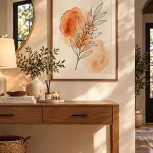 May include: A framed wall art print featuring a black line drawing of an olive branch with leaves and small olives, layered over two soft, watercolor-style orange circles on a cream background, enclosed in a simple light brown wooden frame.