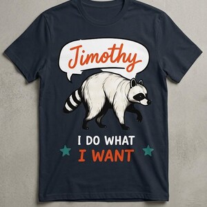 May include: A dark navy blue t-shirt featuring a graphic of a white raccoon with a black mask and striped tail. Above the raccoon is a white speech bubble with the orange text Timothy. Below the raccoon is the white and orange text I DO WHAT I WANT.