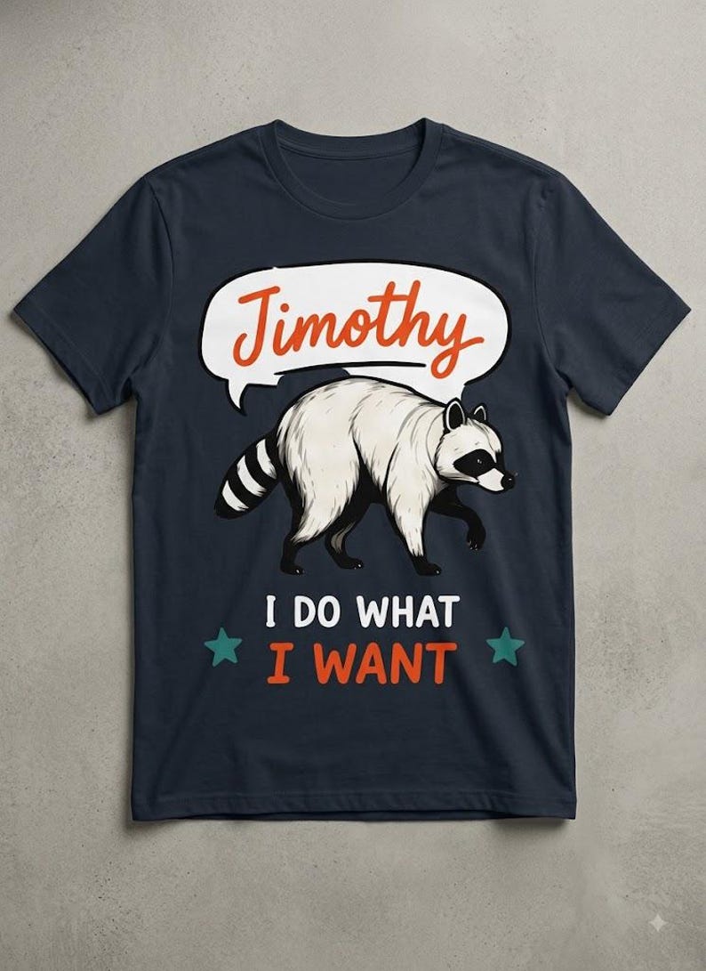 May include: A dark navy blue t-shirt featuring a graphic of a white raccoon with a black mask and striped tail. Above the raccoon is a white speech bubble with the orange text Timothy. Below the raccoon is the white and orange text I DO WHAT I WANT.