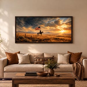 May include: A rectangular landscape wall art print in a dark wooden frame depicting a rider on horseback carrying an American flag across a golden field at sunset with mountains in the distance, displayed above a beige sofa with decorative pillows.