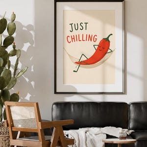 May include: A framed wall art print featuring a cartoon red chili pepper with a smiling face, resting in a beige hammock. The text "JUST CHILLING" is printed in dark green and red letters above the chili pepper on a light cream background.