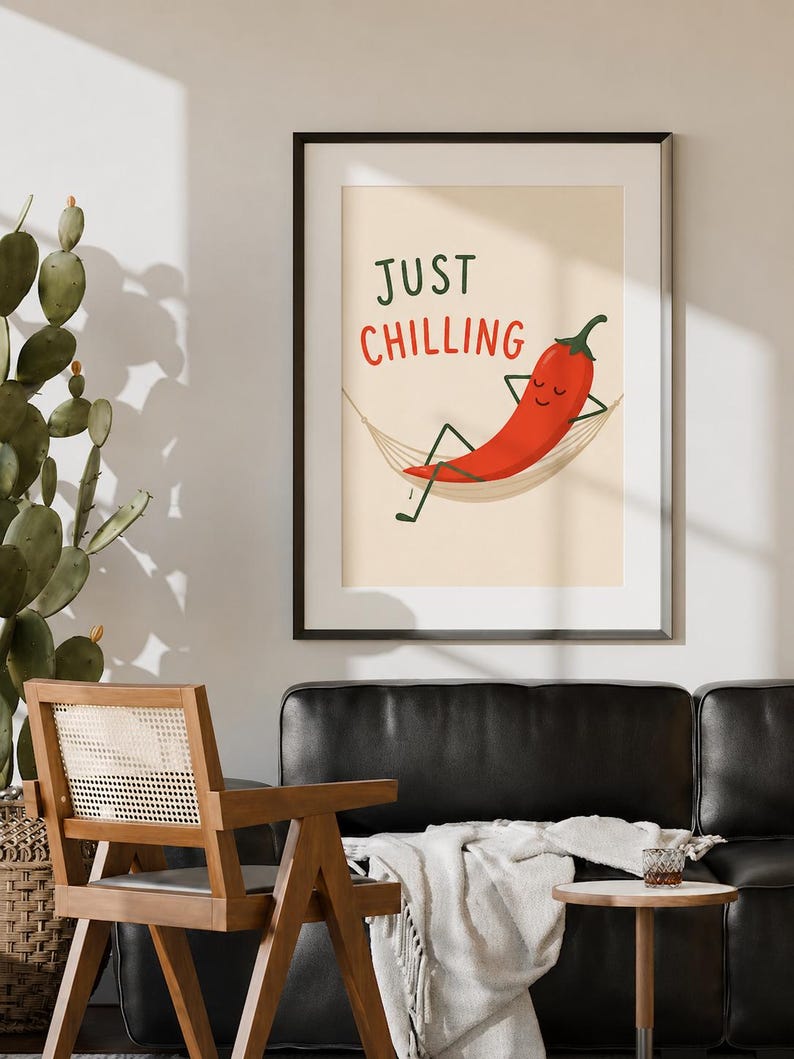May include: A framed wall art print featuring a cartoon red chili pepper with a smiling face, resting in a beige hammock. The text "JUST CHILLING" is printed in dark green and red letters above the chili pepper on a light cream background.