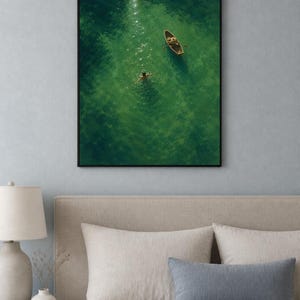 May include: A framed wall art print featuring an aerial view of a small wooden rowboat floating on vibrant green water. A person is swimming in the clear, sunlit water near the boat, creating gentle ripples. The artwork is enclosed in a black frame.