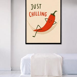 May include: A framed wall art print featuring a cartoon red chili pepper with a smiling face relaxing in a beige hammock. The text "JUST CHILLING" is printed in dark green and red letters above the chili pepper on a light cream background.