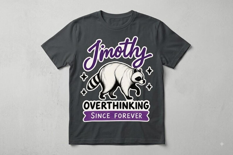 May include: A charcoal grey short-sleeve t-shirt featuring a graphic of a white raccoon with black markings. The text reads "Jimothy" in purple, "OVERTHINKING" in black, and "SINCE FOREVER" in white on a purple banner, accented by four-pointed stars.