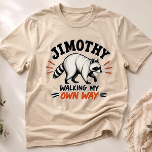 May include: A beige short-sleeve t-shirt featuring a graphic of a raccoon in the centre. Above the raccoon, the text reads JIMOTHY in black. Below the raccoon, the text reads WALKING MY OWN WAY in black and orange with small orange dash accents.
