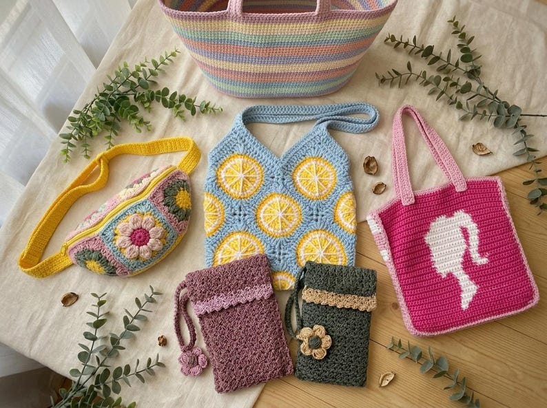 May include: A collection of handmade crochet bags including a large pastel striped tote, a yellow fanny pack with floral motifs, a light blue lemon-patterned shoulder bag, a bright pink tote with a white silhouette, and two small pouches.
