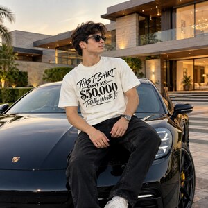 May include: An off-white short-sleeve t-shirt featuring black text that reads "This T-Shirt Cost Me $50,000 Totally Worth It" in a stylised font. The shirt is worn with dark-coloured trousers, a wristwatch, and dark-tinted sunglasses.