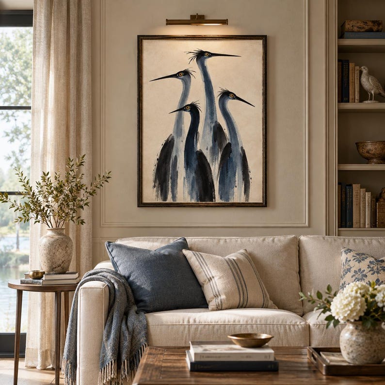 May include: A framed wall art print featuring a stylised watercolour painting of four blue herons with long necks and sharp beaks in shades of dark blue and grey against a neutral beige background, displayed above a beige sofa with throw pillows.