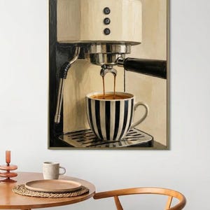 May include: A framed wall art print depicting a cream-colored espresso machine brewing coffee into a black and white striped ceramic mug. The machine features three black circular buttons on the front and a metallic drip tray base.