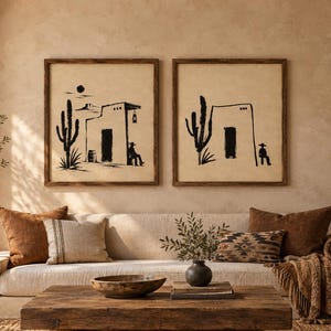 May include: Two square wall art prints in dark wooden frames featuring black ink illustrations of desert scenes with cacti, adobe buildings, and figures in cowboy hats on a light beige background, displayed above a neutral-toned sofa.