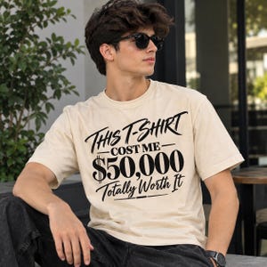 May include: A cream-coloured short-sleeve t-shirt featuring black text that reads: This T-SHIRT COST ME $50,000 Totally Worth It. The shirt has a crew neckline and a relaxed fit, with the graphic printed in a mix of script and bold serif fonts.