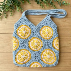 May include: A light blue crochet tote bag featuring a repeating pattern of yellow and white lemon slice motifs. The bag is constructed from joined square panels with a single strap handle, showcasing a textured yarn finish throughout the design.
