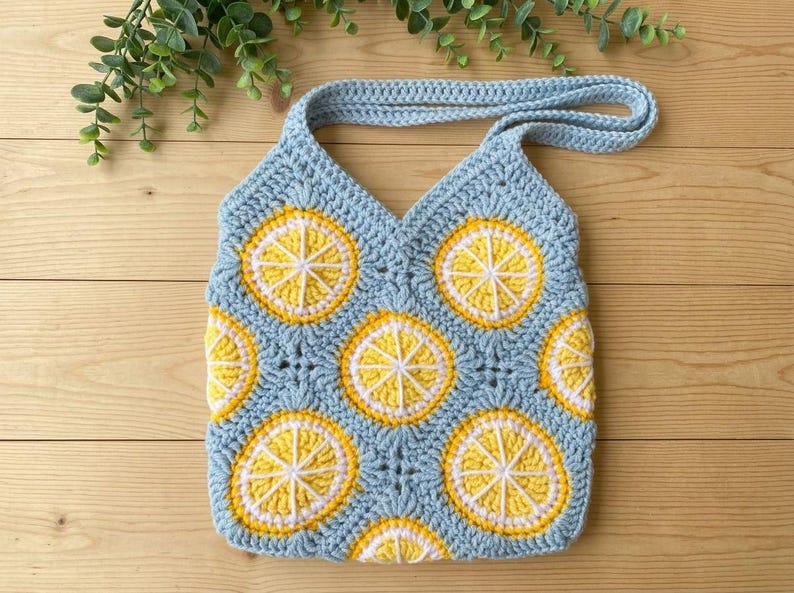May include: A light blue crochet tote bag featuring a repeating pattern of yellow and white lemon slice motifs. The bag is constructed from joined square panels with a single strap handle, showcasing a textured yarn finish throughout the design.