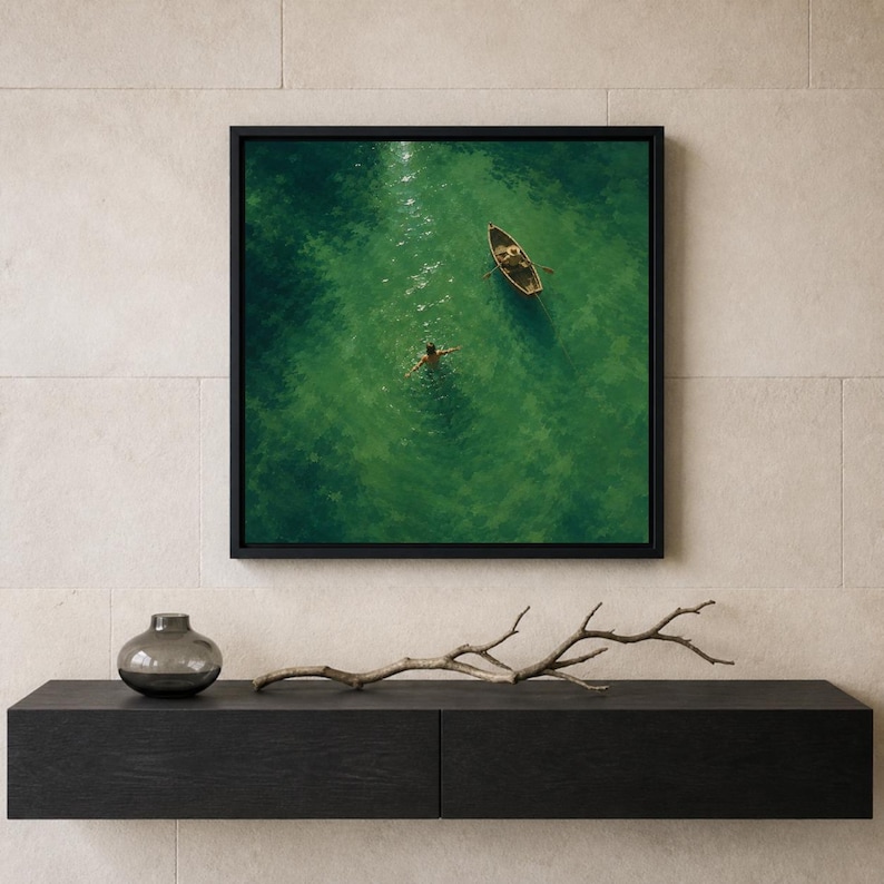 May include: A square framed wall art print depicting an aerial view of a small wooden rowboat floating on vibrant green water next to a person swimming with arms outstretched. The artwork is encased in a thin black frame and mounted on a wall.