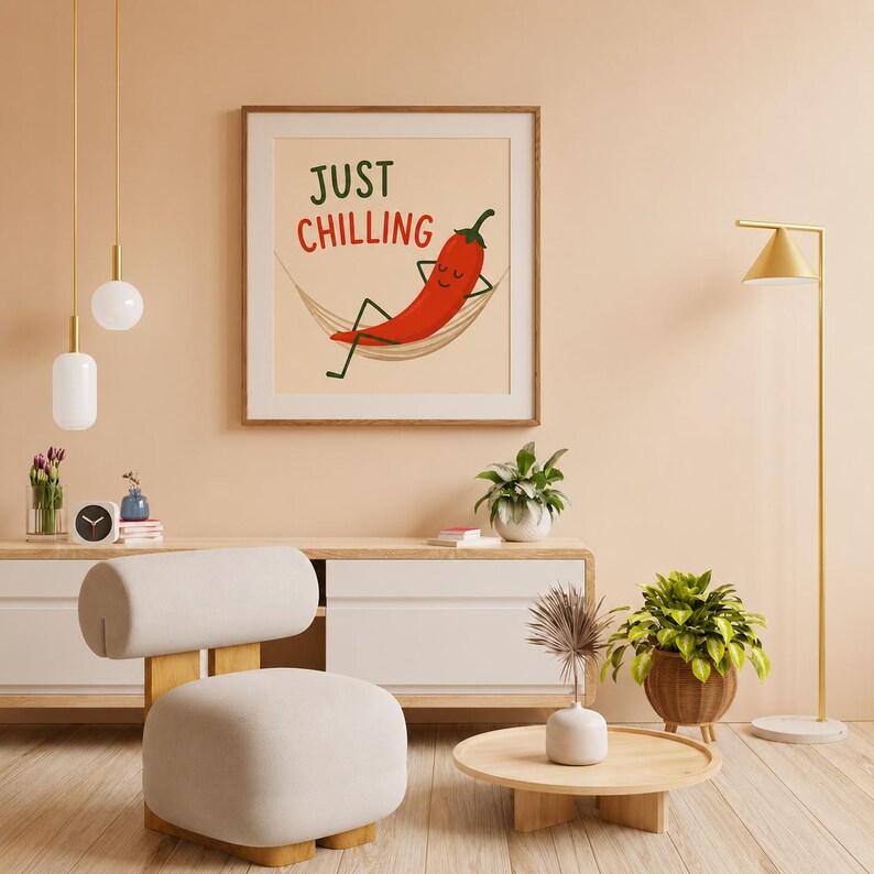 May include: A square framed wall art print featuring a cartoon red chili pepper with a smiling face relaxing in a beige hammock. The text above the chili reads JUST CHILLING in green and red capital letters against a light cream background.