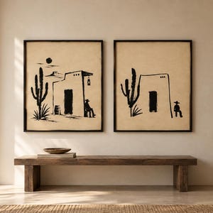 May include: Two framed wall art prints featuring black ink illustrations on a beige background. The left print depicts a desert scene with a cactus, a building, a sun, and a seated figure. The right print shows a similar building and cactus.