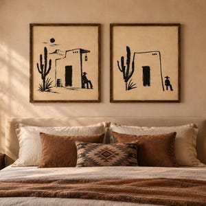 May include: Two square framed wall art prints featuring black ink illustrations on a beige background. The artwork depicts desert scenes with saguaro cacti, adobe-style buildings, and figures of people sitting, all enclosed in dark wooden frames.
