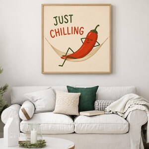 May include: A square wall art print in a light wood frame featuring a cartoon red chili pepper relaxing in a beige hammock. The text "JUST CHILLING" is printed above the chili pepper in dark green and red lettering on a cream-colored background.