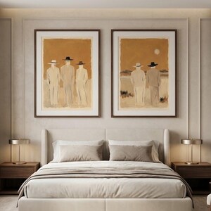 May include: Two framed wall art prints displayed above a bed. The left print shows three figures in hats against a tan background. The right print depicts two figures in hats standing in a landscape under a pale sun. Both feature brown wooden frames.