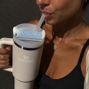 Può includere: Un bicchiere Stanley color crema con manico e cannuccia, dotato di un'etichetta rettangolare azzurra fissata sul coperchio con il nome Amanda stampato in corsivo blu scuro. Il logo Stanley è impresso in rilievo sul lato del bicchiere.