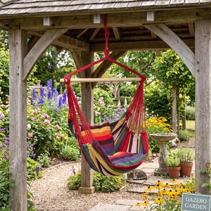 May include: A hanging chair featuring a multi-coloured striped fabric seat in shades of red, orange, yellow, purple, and green. The chair is suspended by red ropes attached to a horizontal wooden spreader bar, creating a comfortable seat.