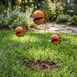Könnte beinhalten: Drei rostfarbene Metall-Gartenstecker in Form von fleischfressenden Pflanzen mit kugelförmigen Köpfen, gezackten Zähnen und herausragenden Zungen. Jede Pflanze hat einen runden Körper mit blütenblattartigen Rückseiten auf dünnen Metallstielen.