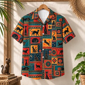 Puede incluir: Una camisa de manga corta con botones que presenta un vibrante patrón de patchwork de siluetas de gatos negros, formas geométricas y estrellas en tonos verde azulado, naranja y rojo. La camisa tiene un cuello clásico y botones blancos.