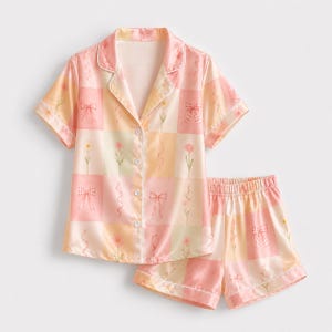 Peut inclure: Un ensemble de pyjama composé d'une chemise à manches courtes boutonnée et d'un short assorti, présentant un motif à carreaux dans des tons doux de rose, jaune et crème. Le tissu est orné de petites illustrations de fleurs et de rubans.