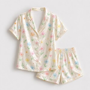 May include: A cream-colored pajama set consisting of a short-sleeved button-down shirt with a notched collar and matching elastic-waist shorts. The fabric features a repeating hand-drawn style floral pattern in shades of blue, pink, orange, and green.