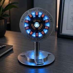 May include: A circular metallic arc reactor replica mounted on a cylindrical stand and base. The center glows with a bright blue light, surrounded by copper-colored coils. The outer ring features the engraved text: PROOF THAT TONY STARK HAS A HEART.
