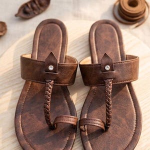 May include: A pair of brown flat sandals featuring a textured finish, a braided T-strap design, and a single toe loop. Each sandal includes a wide horizontal strap across the instep adorned with a small, centered, clear circular rhinestone accent.