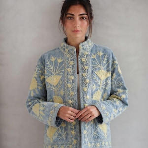 Può includere: Una giacca azzurra con colletto alto e maniche lunghe, decorata con intricati ricami floreali gialli sul davanti e sulle maniche. Il capo presenta un orlo dritto e una chiusura frontale con bottoni in tessuto coordinati.