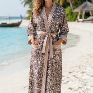 May include: A knee-length robe featuring a brown and black leopard print pattern. The garment includes a beige and white vertical striped trim along the front opening, sleeve cuffs, pockets, and the matching tie belt at the waist.