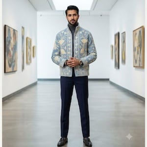 May include: A light blue jacket featuring intricate cream-colored Kashmiri embroidery, a high collar, and a front zipper closure. The garment is worn over a grey turtleneck sweater, paired with dark navy trousers and dark brown leather loafers.